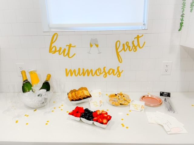 Brunch spread on white kitchen counter with gold "but first mimosas" sign, ice bucket of champagne and orange juice, fresh berries, sliced bread and pastries.