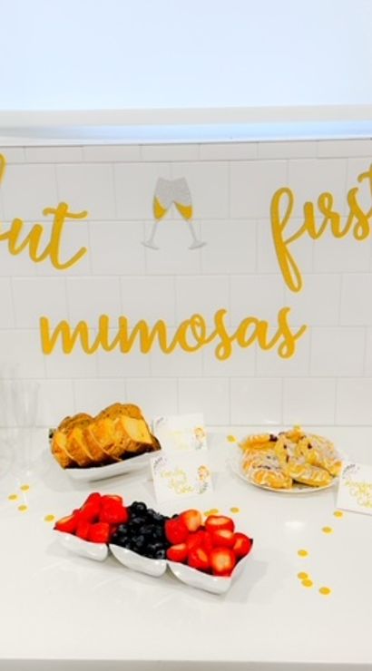 Brunch spread on white kitchen counter with gold "but first mimosas" sign, ice bucket of champagne and orange juice, fresh berries, sliced bread and pastries.