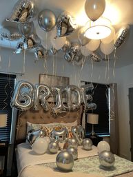 Bedroom decorated for a bridal celebration with silver and white helium balloons on the ceiling, large silver foil letters spelling "BRIDE" above the bed, and silver heart and round balloons scattered on the bed.
