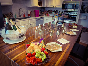 Festive home kitchen island set for a 40th birthday: champagne bottles on ice, gold plastic flutes, metallic party hats and confetti, charcuterie platter with strawberries, grapes, crackers and cheese, and "Happy 40th birthday" plates and napkins with white cabinets and stainless appliances in the background.