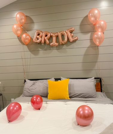 Bachelorette bedroom decor with rose-gold "BRIDE" foil letter balloons and matching balloons above a bed with gray bedding, bright yellow accent pillow, and rose-gold balloons scattered on a white comforter.