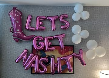 Indoor patterned wall decorated with metallic pink letter balloons reading "LETS GET MASHITTY", a large pink cowboy boot foil balloon on the left, white cluster balloons on the right, and an abstract framed painting below.