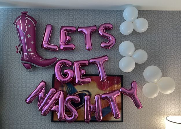 Indoor patterned wall decorated with metallic pink letter balloons reading "LETS GET MASHITTY", a large pink cowboy boot foil balloon on the left, white cluster balloons on the right, and an abstract framed painting below.