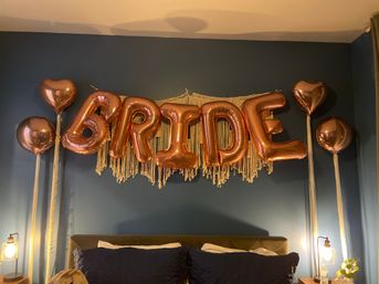 Indoor bridal bedroom decor: rose gold foil balloons spelling BRIDE with heart balloons and macramé fringe above a bed against a navy blue wall, bedside lamps and pillows.