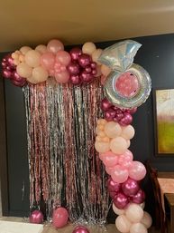 Indoor pink and rose-gold balloon garland framing a shimmering rose-gold fringe curtain, topped by a sparkly diamond-ring foil balloon — playful engagement or bridal-shower photo backdrop.