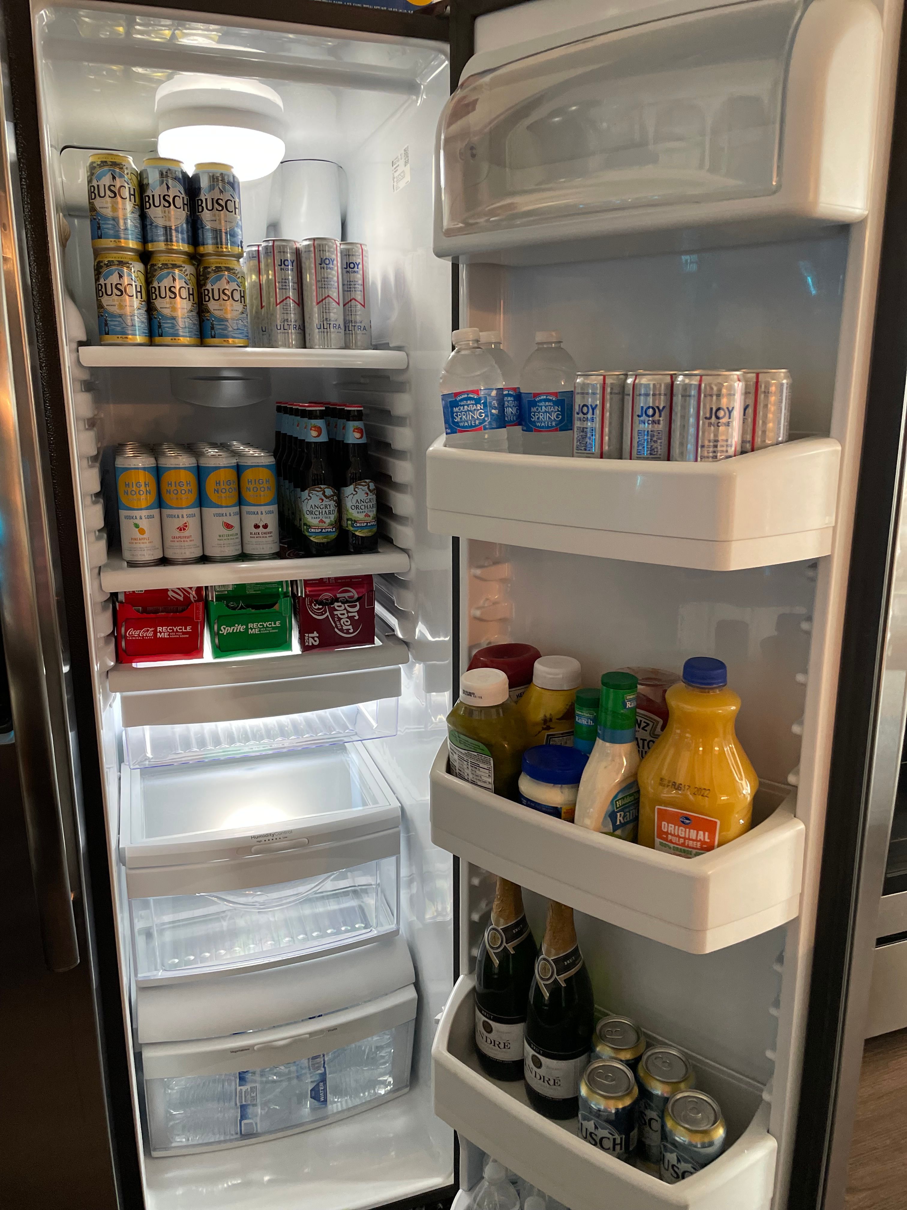 Open home refrigerator interior showing organized chilled beverages: stacked cans of beer and seltzer, bottled water, mixers and condiments, orange juice and champagne on door shelves.