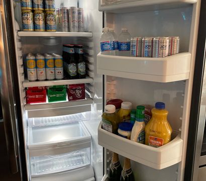 Open home refrigerator interior showing organized chilled beverages: stacked cans of beer and seltzer, bottled water, mixers and condiments, orange juice and champagne on door shelves.
