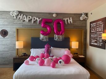 Hotel room bed decorated for a 50th birthday: silver 'HAPPY' letters and large pink '50' balloons with tassels above a bed scattered with pink and black‑spotted balloons, bedside lamps, a cowboy hat on the wall and Nashville-themed wall art.