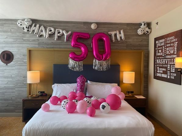 Hotel room bed decorated for a 50th birthday: silver 'HAPPY' letters and large pink '50' balloons with tassels above a bed scattered with pink and black‑spotted balloons, bedside lamps, a cowboy hat on the wall and Nashville-themed wall art.