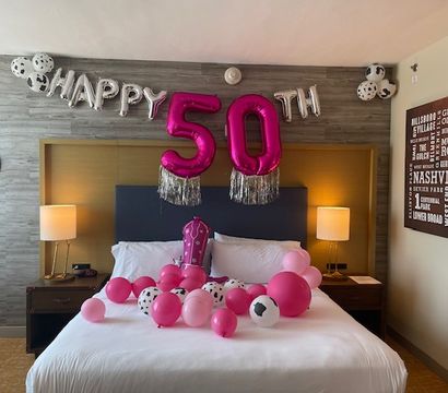 Hotel room bed decorated for a 50th birthday: silver 'HAPPY' letters and large pink '50' balloons with tassels above a bed scattered with pink and black‑spotted balloons, bedside lamps, a cowboy hat on the wall and Nashville-themed wall art.