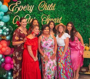Seven smiling women in colorful dresses pose together in front of a leafy green backdrop with a balloon garland and gold script reading "Every Child" at a festive, celebratory event