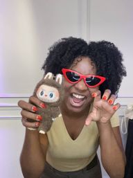Playful person with curly hair in red cat-eye sunglasses and red nails holding a brown plush toy, smiling and posing indoors.