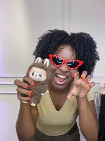 Playful person with curly hair in red cat-eye sunglasses and red nails holding a brown plush toy, smiling and posing indoors.