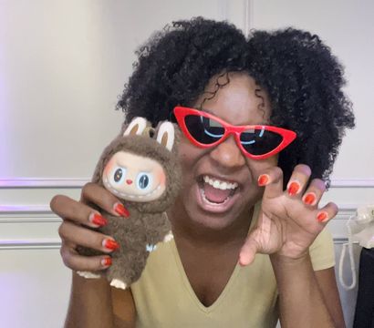 Playful person with curly hair in red cat-eye sunglasses and red nails holding a brown plush toy, smiling and posing indoors.