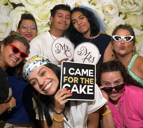 Group of seven in a wedding photo booth with white rose backdrop — bride wearing a veil holding 'Mr' and 'Mrs' signs, friend smiling front-center holding an 'I CAME FOR THE CAKE' prop, others in playful sunglasses.