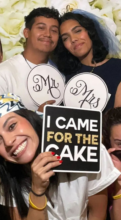 Group of seven in a wedding photo booth with white rose backdrop — bride wearing a veil holding 'Mr' and 'Mrs' signs, friend smiling front-center holding an 'I CAME FOR THE CAKE' prop, others in playful sunglasses.
