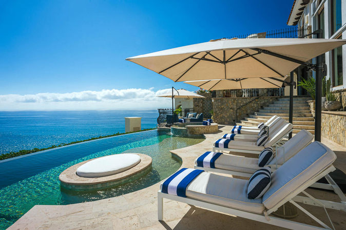 Oceanfront infinity pool at a coastal villa with a circular sunpad, stone terrace, blue-and-white striped cushioned lounge chairs and large umbrellas overlooking the sparkling sea and clear sky.