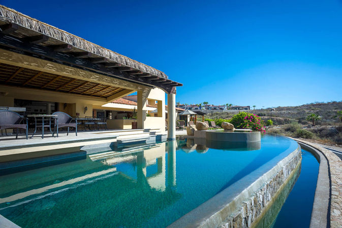 Infinity pool at a luxury villa with thatched pergola, outdoor dining area, sun loungers and arid hillside under a bright blue sky