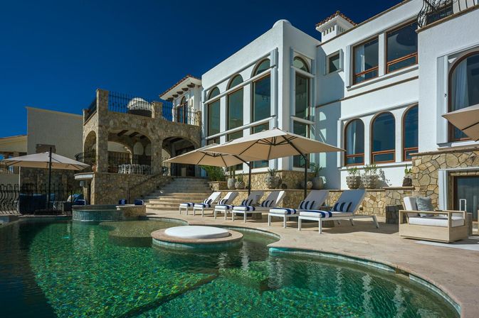 Luxury Mediterranean-style villa with stone terrace and arched windows, turquoise swimming pool featuring a round sun platform, striped lounge chairs and umbrellas under a clear blue sky.