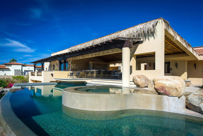 Luxury coastal villa with a thatched-roof terrace, outdoor bar seating and an infinity pool with built-in spa under a bright blue sky