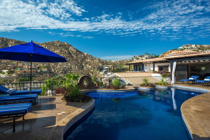 Sunny luxury hillside pool terrace with blue-striped chaise lounges and umbrella, Mediterranean-style villa, rocky hills and bright blue sky.