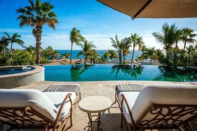 Oceanfront infinity pool and spa at a tropical beachfront resort, palm trees framing a sparkling blue ocean horizon, two cushioned sun loungers and a small table under an umbrella.