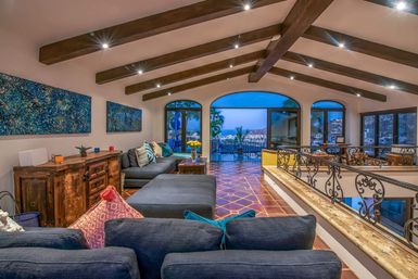 Open-concept Mediterranean-style living room with exposed wooden beams, terracotta tile floor, deep-blue sectional, wrought-iron railing and arched glass doors framing a coastal bay dusk view.