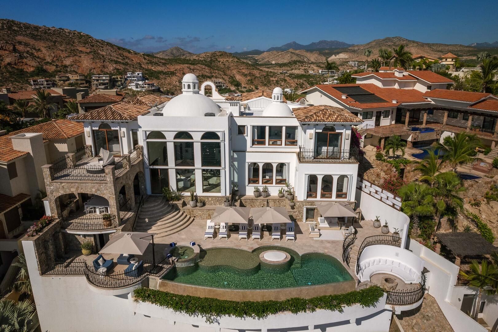 Sun-soaked luxury Mediterranean-style villa perched on a hillside with white domes, multi-level stone terraces, palm trees, sun loungers, and a curved infinity pool overlooking arid mountain scenery.