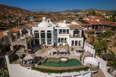 Sun-soaked luxury Mediterranean-style villa perched on a hillside with white domes, multi-level stone terraces, palm trees, sun loungers, and a curved infinity pool overlooking arid mountain scenery.