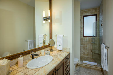 Bright, spa-like bathroom with stone-tile vanity and floor, oval white sink with dark wood cabinets, large framed mirror and wall sconce, folded white towels, and a glass-enclosed walk-in shower with frosted window.