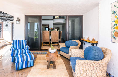 Sunlit covered patio lounge with a bold blue-striped armchair, two woven rattan chairs with round blue cushions, a rustic wooden coffee table on a jute rug, decorative tile trim and sliding glass doors opening to a dining room.