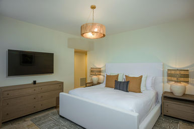 Contemporary neutral-toned master bedroom interior with white upholstered bed, amber accent pillows, matching wood dressers, woven pendant light and wall-mounted TV.
