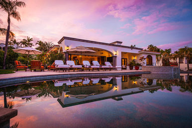 Tropical luxury villa patio with sun loungers and umbrellas along a reflecting pool at sunset, palm trees framing a pink and purple sky.