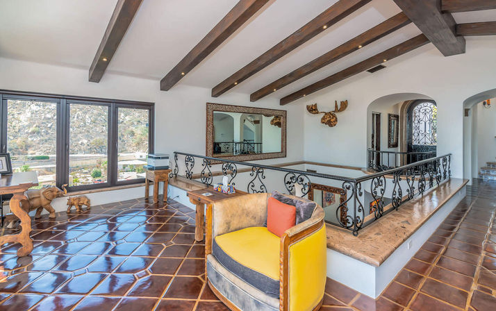 Sunlit Mediterranean-style loft with exposed wood beams, glossy terracotta tile, ornate wrought-iron railing, panoramic hillside windows and a cheerful yellow accent chair.