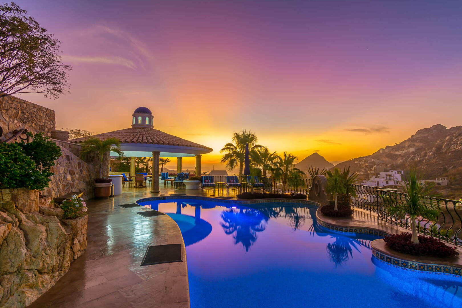 Sunset at an oceanview luxury resort pool — blue-lit curved infinity pool reflecting palm trees, a tiled gazebo, and rocky cliffs under a purple-orange sky.