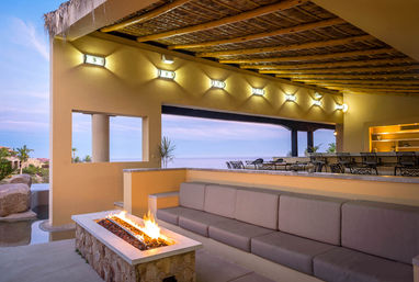 Cozy oceanfront resort patio with woven palapa ceiling, built-in cushioned sofa, stone fire pit, bar seating and sunset sea view