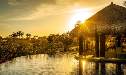 Golden sunset over a tropical infinity pool with a thatched palapa bar, silhouetted palm trees and shimmering reflective water