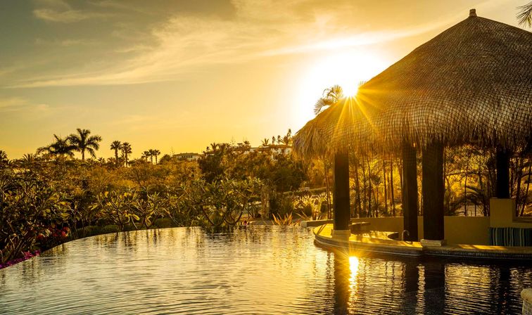 Golden sunset over a tropical infinity pool with a thatched palapa bar, silhouetted palm trees and shimmering reflective water