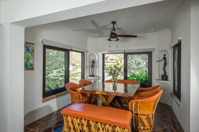 Sunlit Mexican-style dining room with orange leather chairs and a wooden table, tiled floor, ceiling fan, and large windows overlooking hillside homes and lush greenery.