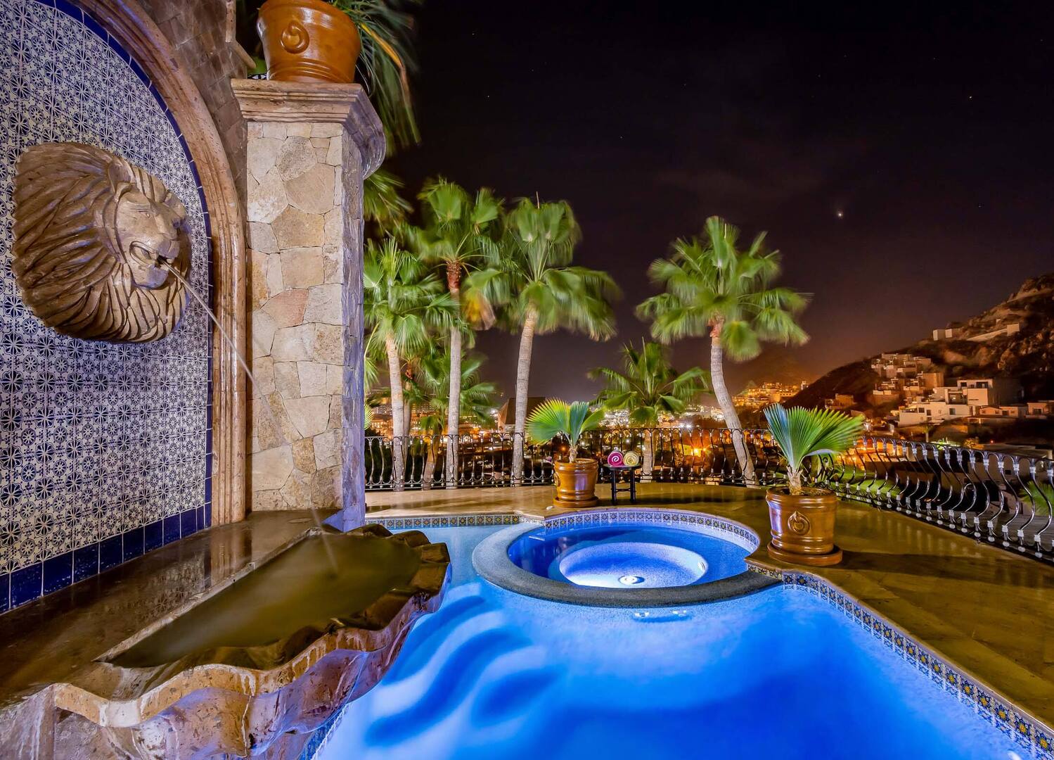 Blue-lit luxury pool and circular hot tub on a coastal hillside terrace at night, palm trees, decorative tiled wall with lion-head fountain, potted palms and glowing city lights below.