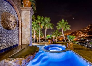 Blue-lit luxury pool and circular hot tub on a coastal hillside terrace at night, palm trees, decorative tiled wall with lion-head fountain, potted palms and glowing city lights below.