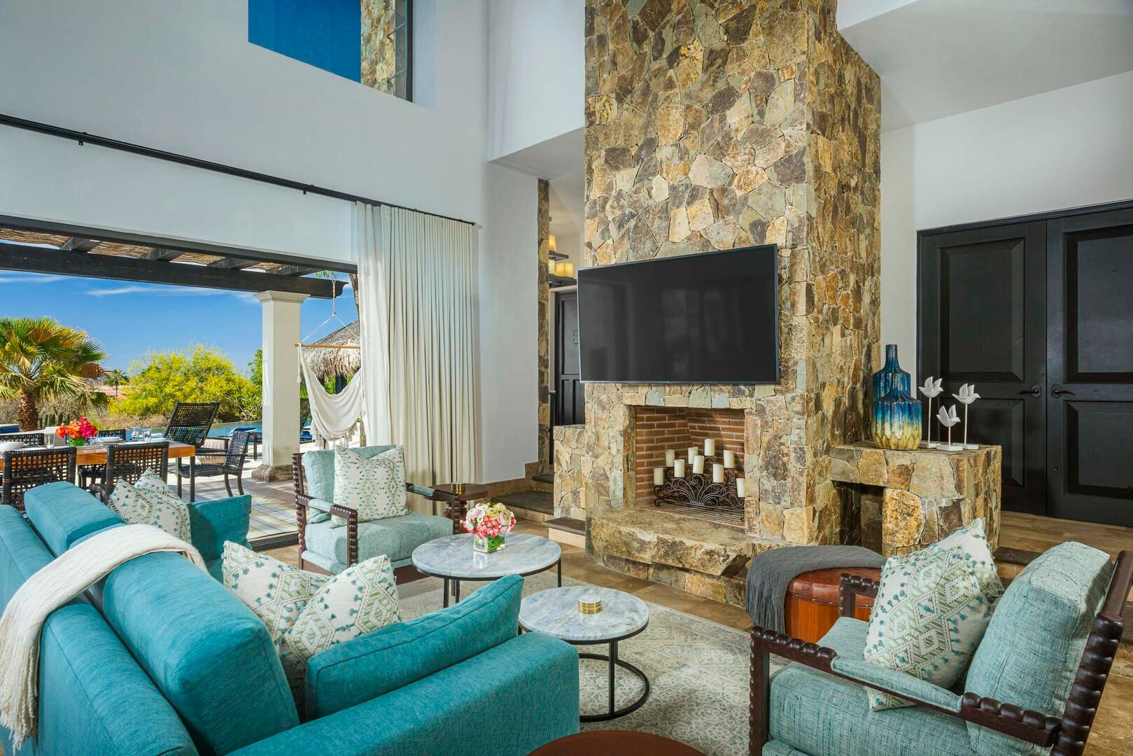 Bright open-plan coastal villa living room with turquoise sofas, stone fireplace and wall-mounted TV opening onto a sunny pool terrace with hammock and tropical plants.