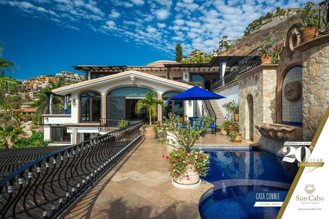 Sunny luxury villa terrace in Los Cabos, Mexico with a tiled plunge pool, blue patio umbrella, stone lion fountain, arched glass doors, potted palms and hillside homes under a bright blue sky.