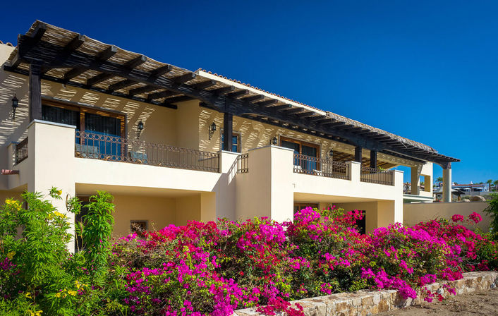 Sunny Mediterranean-style coastal villa with dark wooden pergola, second-floor balconies, and vibrant pink bougainvillea in front under a deep blue sky.