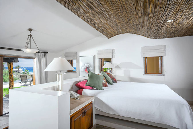 Bright beachfront bedroom with a large white bed topped with colorful pillows, woven wood ceiling, bedside lamps and sliding doors opening to a patio and ocean view