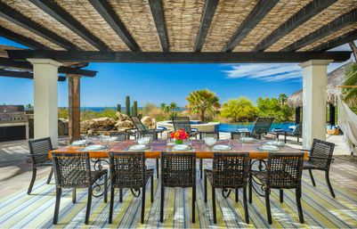 Oceanfront outdoor dining patio under a woven pergola — long wooden table set for eight, pool and lounge chairs, palm trees and cacti in a sunny coastal setting.