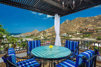 Sunny coastal terrace with striped blue patio chairs around a round glass table holding a yellow flower, shaded by a pergola and overlooking palm trees, a seaside town and rocky coastal hills under a bright blue sky.