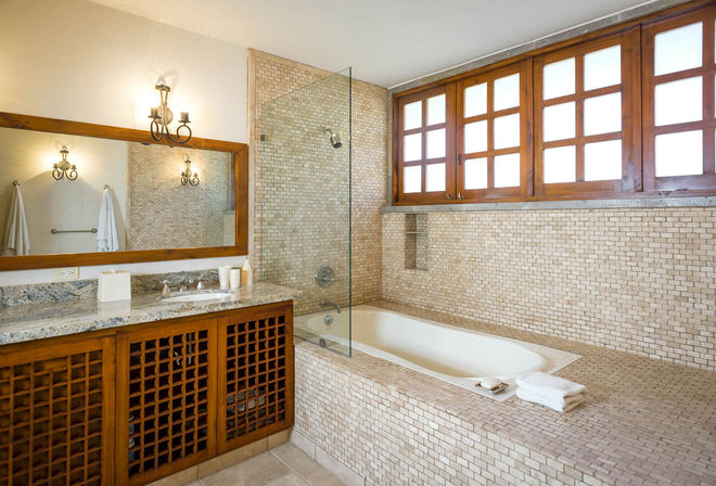 Sunlit spa-like beige mosaic-tiled bathroom with built-in soaking tub, glass shower screen, granite vanity, and wood-framed windows.