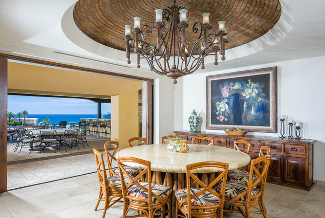 Inviting coastal dining room with a round marble table surrounded by rattan chairs with tropical cushions, wrought-iron chandelier beneath a brick dome, wooden sideboard with floral painting, and open patio leading to ocean-view terrace seating.