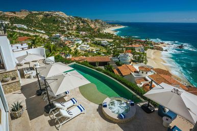 Sun-soaked cliffside oceanfront terrace of a luxury villa with an infinity pool, bubbling hot tub, striped lounge chairs and umbrellas overlooking a sandy beach, turquoise ocean and coastal hillside homes.